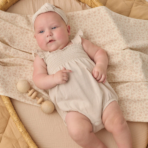 Newborn baby in a beige romper lying on a textured blanket with a wooden rattle.