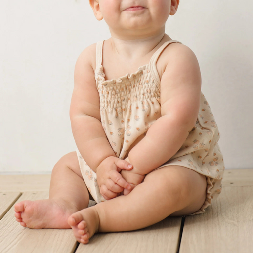 Baby sitting on a wooden floor wearing a beige romper with a white background