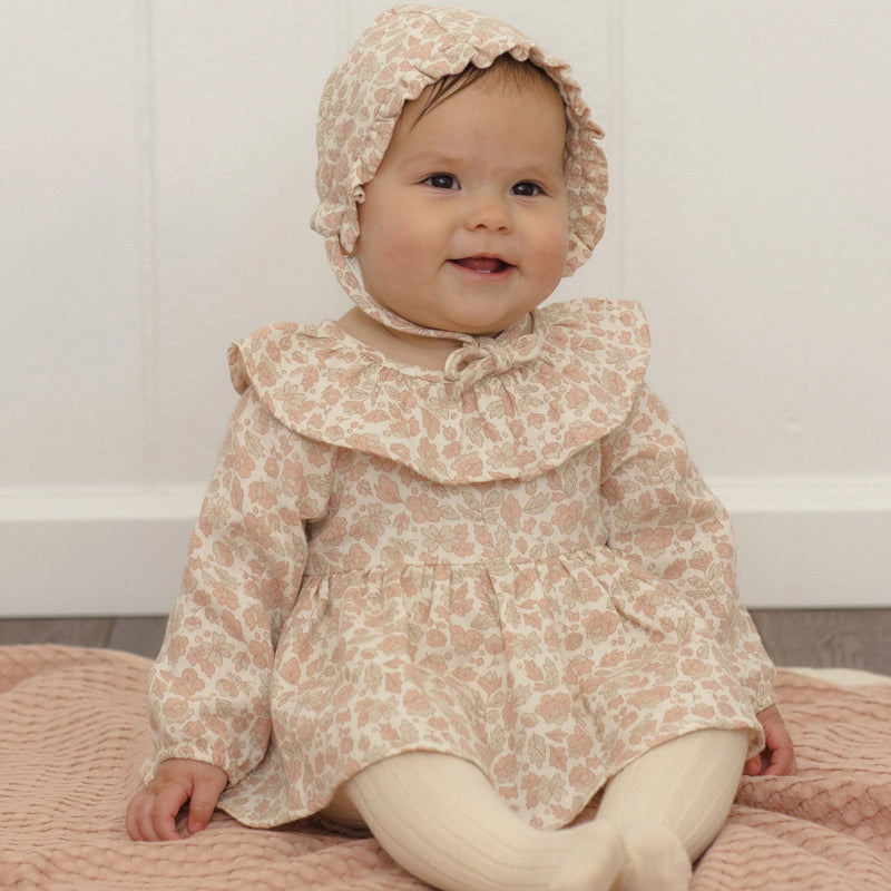 A baby wearing a light beige and pink floral print long-sleeve dress with a large ruffled collar, matching bonnet, and cream tights, sitting on a pink quilted blanket against a white paneled wall.