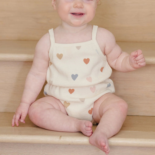 Baby wearing a sleeveless top and shorts with heart patterns sitting on wooden steps.