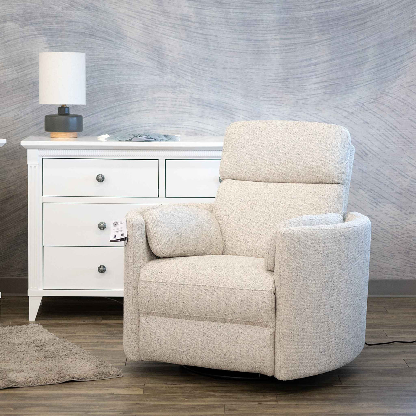 A cream-colored upholstered swivel recliner chair positioned in front of a white three-drawer dresser with a lamp, set against a textured gray wall and wood-look flooring.