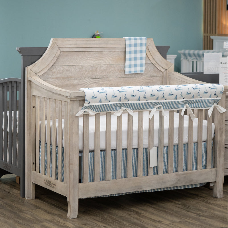 A light natural wood convertible crib with a tall arched headboard and side rails, styled with a blue and white checkered blanket and a blue bird-patterned bumper roll in a nursery with a blue wall and wood floor.