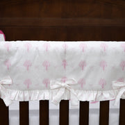 White crib bumper with a pink tree pattern and ruffled trim, tied to the crib slats with white bows, shown in a nursery setting.