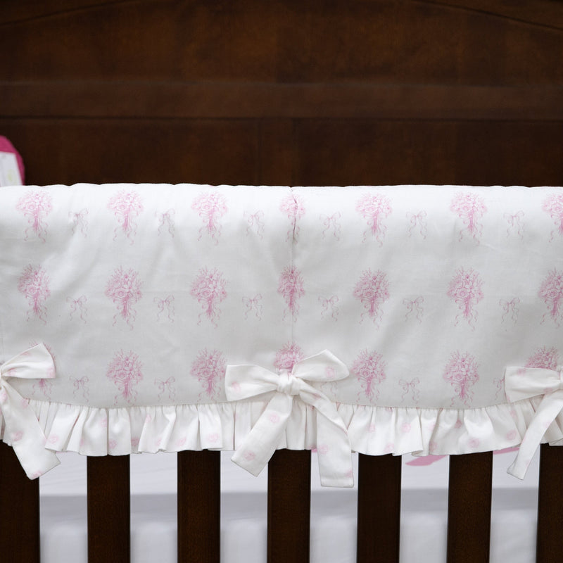 White crib bumper with a pink tree pattern and ruffled trim, tied to the crib slats with white bows, shown in a nursery setting.
