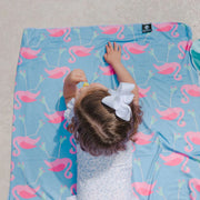 Child playing on a blue mat with pink flamingo pattern, wearing a white dress with a bow.
