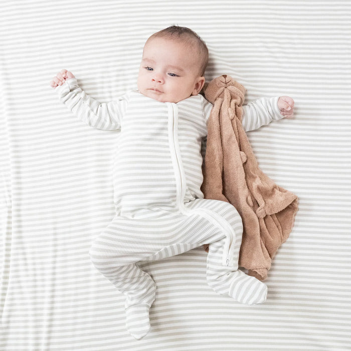 A baby lying on a matching striped sheet wearing the footie while holding a plush lovey.