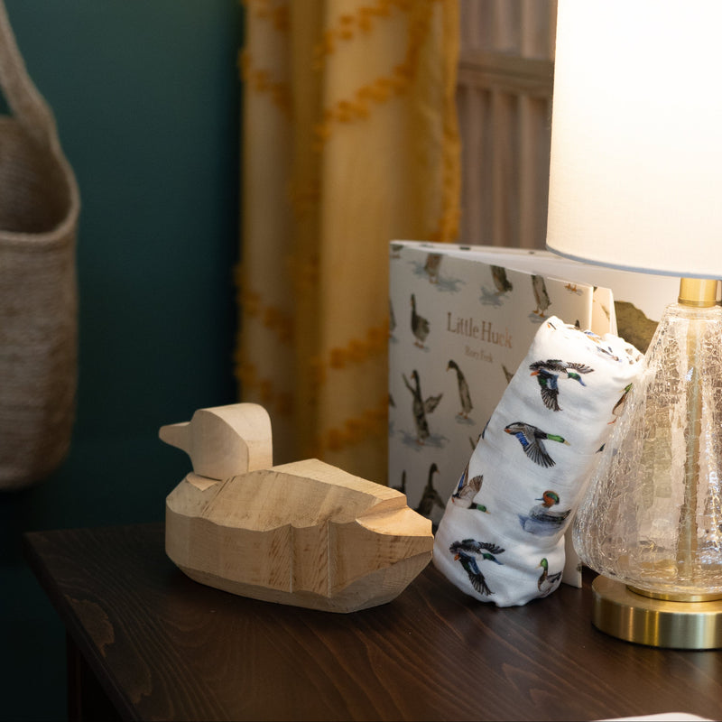 Wooden duck figurine, lamp, and ducjk patterned swaddle blanket on a table with curtains in the background.