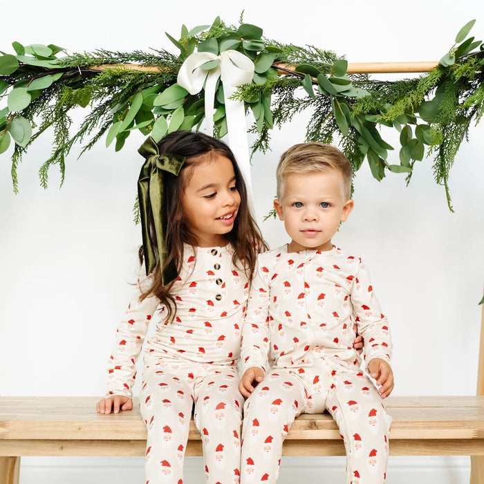 Two children in matching pajamas sitting on a wooden bench with a festive greenery backdrop.