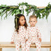 Two children in matching pajamas sitting on a wooden bench with a festive greenery backdrop.