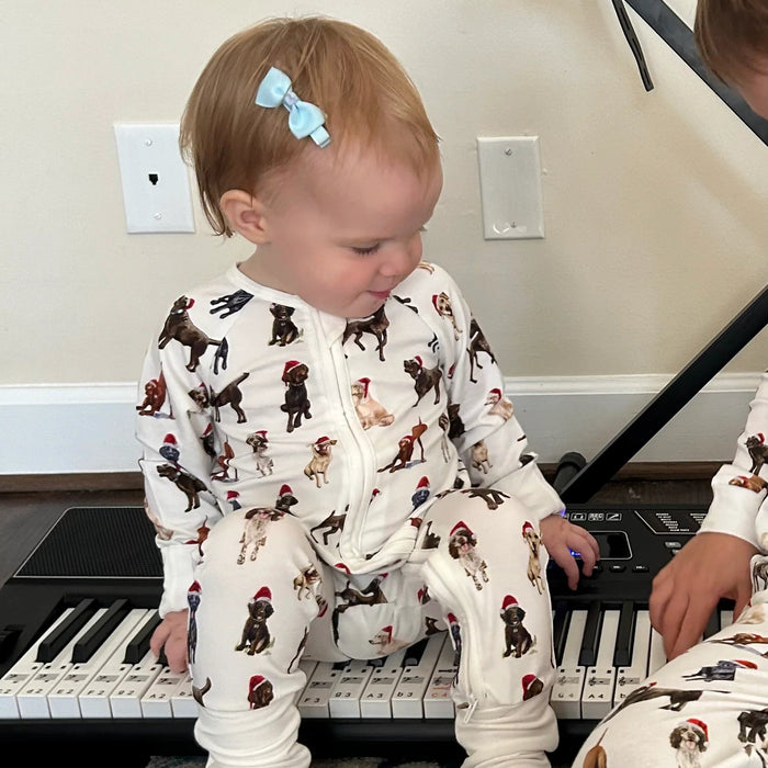 Toddler wearing Saltwater Swaddles Santa Dogs footie with holiday dog print while sitting indoors beside a piano