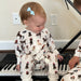 Toddler wearing Saltwater Swaddles Santa Dogs footie with holiday dog print while sitting indoors beside a piano
