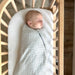 A newborn sleeps peacefully in a crib while wrapped in the Checkmate Sleep Sack Swaddle. The swaddle fits snugly around the baby with the zipper visible along the side.