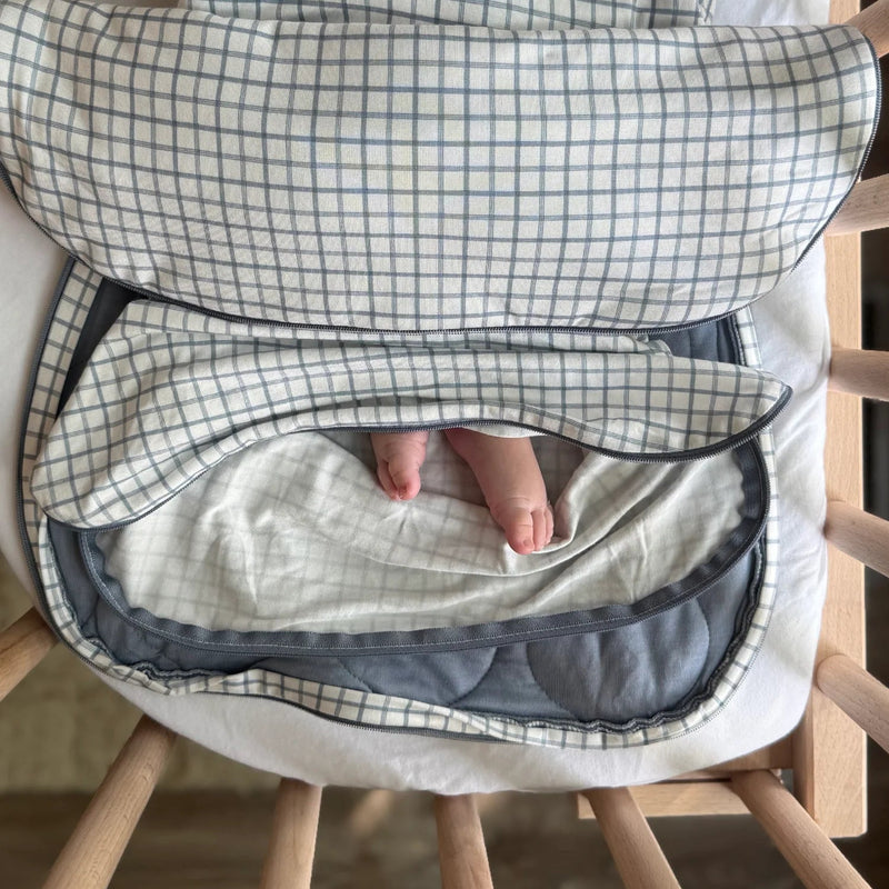 The swaddle is opened to reveal the inner belly band and the soft interior lining. A baby’s legs are visible resting comfortably inside the open swaddle.