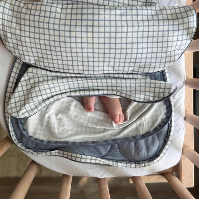 The swaddle is opened to reveal the inner belly band and the soft interior lining. A baby’s legs are visible resting comfortably inside the open swaddle.