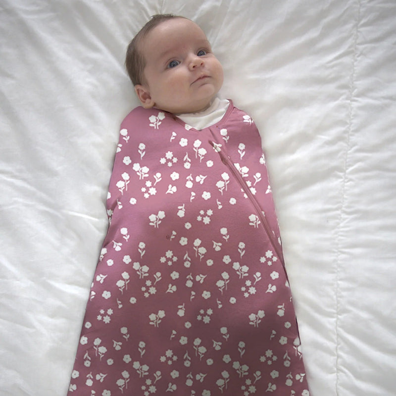 A newborn sleeps on a white surface wearing the Mini Floral Sleep Sack Swaddle fully zipped.