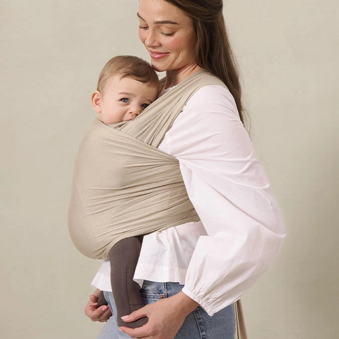 Woman holding a baby in a beige baby carrier against a plain background