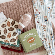 A baby gift set arranged on a beige knit blanket, including a football-themed baby book, a plush football toy, patterned baby clothes, and folded fabric items on a white background.