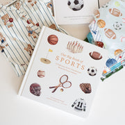 A styled flat-lay of sports-themed stationery and books on a white surface. The set includes a striped paper notepad with teddy bears, a white hardcover book titled 'Tummy Time Sports,' a white hardcover book titled 'Our Big Book of Sports,' and a patterned paper bag or envelope with sports illustrations.