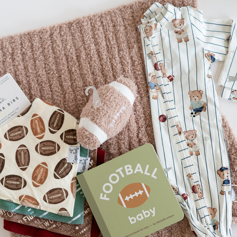 A baby gift set arranged on a beige knit blanket, including a football-themed baby book, a plush football toy, patterned baby clothes, and folded fabric items on a white background.