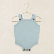 A light blue knit baby bodysuit with shoulder straps and decorative tortoiseshell buttons, hanging on a gold wire hanger against a textured beige background.