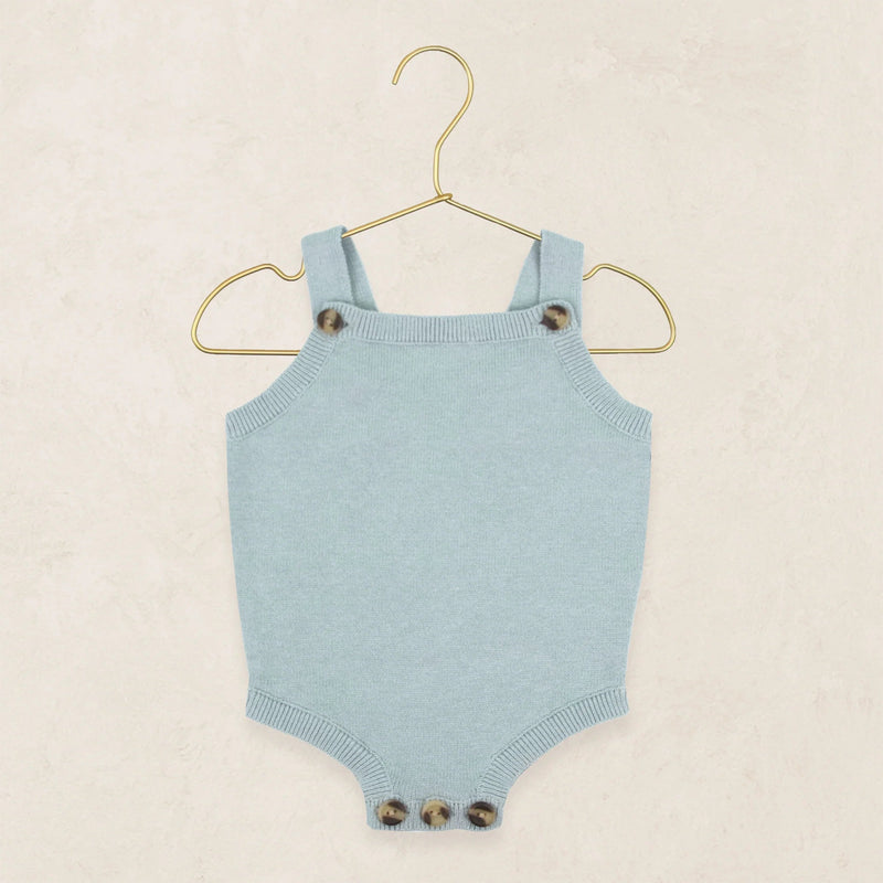 A light blue knit baby bodysuit with shoulder straps and decorative tortoiseshell buttons, hanging on a gold wire hanger against a textured beige background.