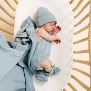 A sleeping newborn baby wearing a light blue knit beanie, matching blue footie, and a light blue blanket, lying on a white mattress inside a wicker bassinet.