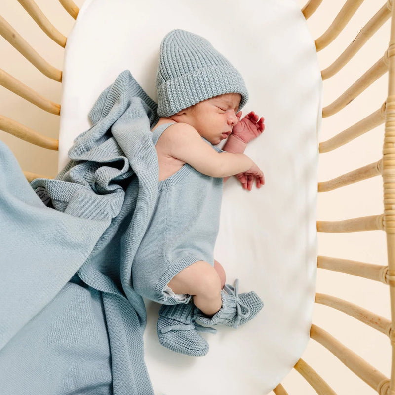 A sleeping newborn baby wearing a light blue knit beanie, matching blue footie, and a light blue blanket, lying on a white mattress inside a wicker bassinet.