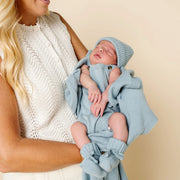 A smiling blonde woman holds a sleeping newborn baby wrapped in a light blue knit blanket and matching hat with booties, against a plain beige studio background.
