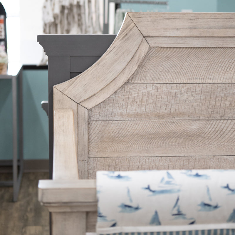 A close-up of a light beige wood-look crib bumper or crib panel with a scalloped top edge, shown in a nursery setting with a blue and white sailboat-patterned crib sheet visible below.