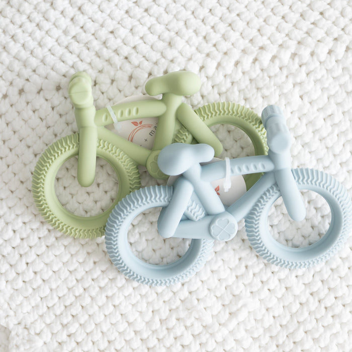 Two bicycle-shaped teething rings, one green and one blue, on a textured white surface.
