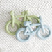 Two bicycle-shaped teething rings, one green and one blue, on a textured white surface.
