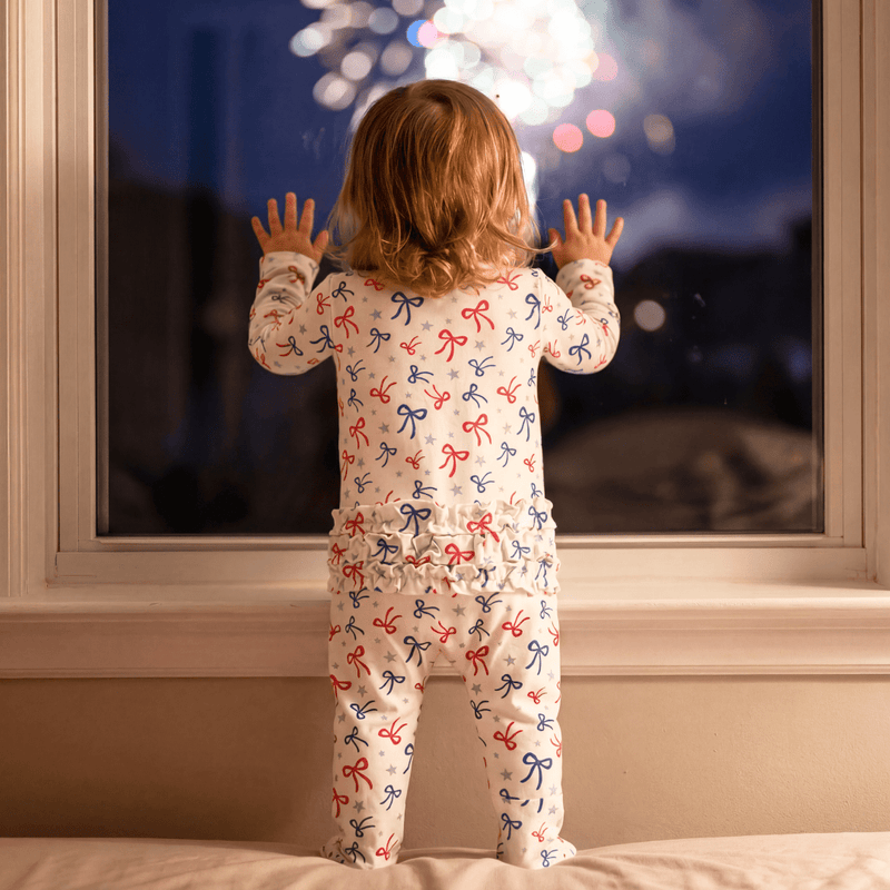 Child in pajamas with bow pattern looking out a window at fireworks