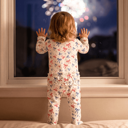 Child in pajamas with bow pattern looking out a window at fireworks