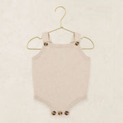 A cream-colored knit baby bodysuit with button details on the shoulder straps and crotch, hanging on a gold wire hanger against a textured beige background.