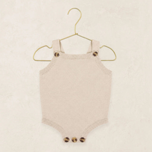 A cream-colored knit baby bodysuit with button details on the shoulder straps and crotch, hanging on a gold wire hanger against a textured beige background.