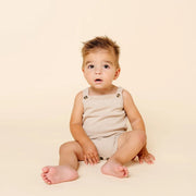 A baby sitting cross-legged on a warm cream studio background, wearing a sleeveless beige romper.