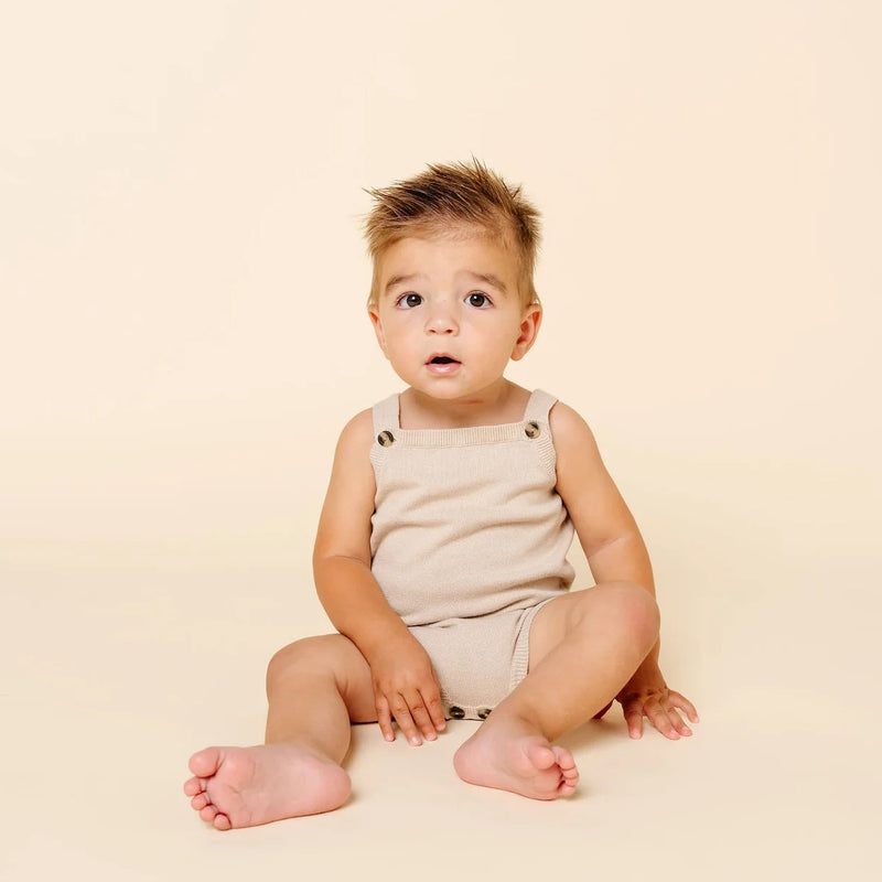 A baby sitting cross-legged on a warm cream studio background, wearing a sleeveless beige romper.