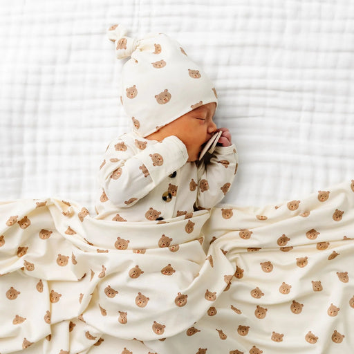 Newborn baby wrapped in a white blanket with brown patterns, wearing a matching hat, on a white textured surface.