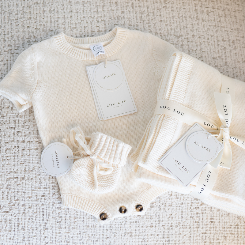 Baby sweater knit onesie, baby booties and blanket set with 'LOU LOU' tags on a textured surface