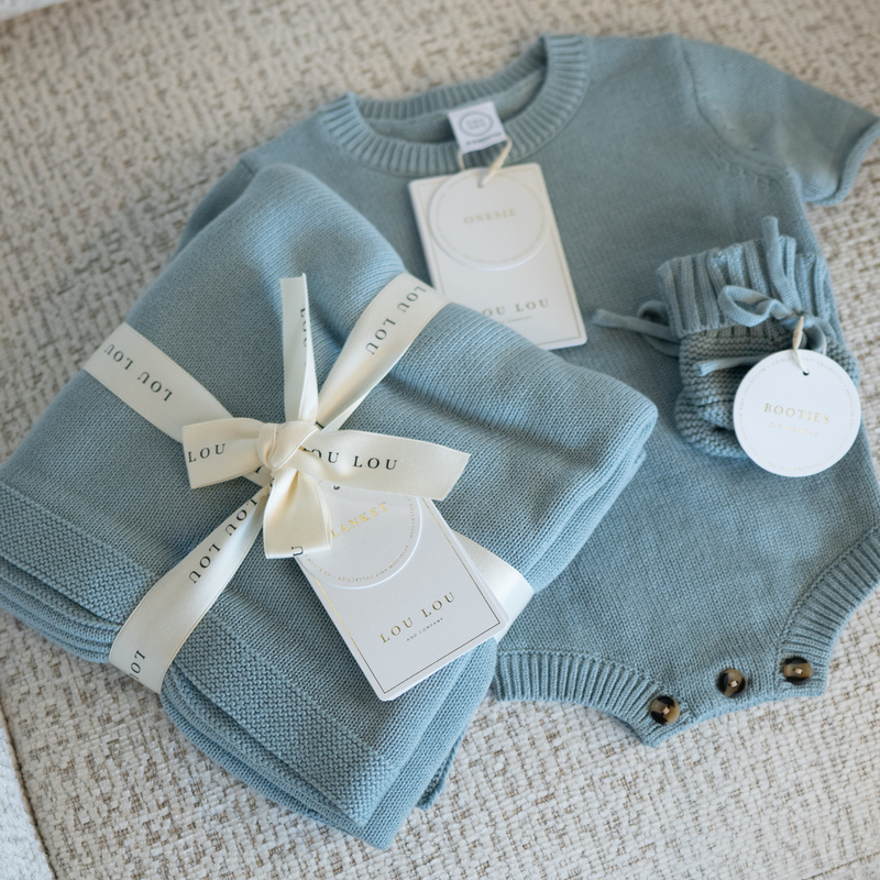 Blue baby knit sweater onesie with matching baby blanket and booties on a textured surface