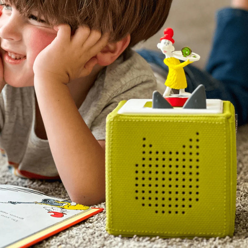 A young boy smiling while reading the Green Eggs and Ham book next to a green Toniebox with the Sam-I-Am figurine on top