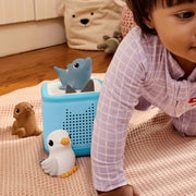 A baby crawling on a pink textured blanket next to a light blue toy basket filled with bath toys, including a blue dolphin, a white duck, and a brown seal in a home setting.