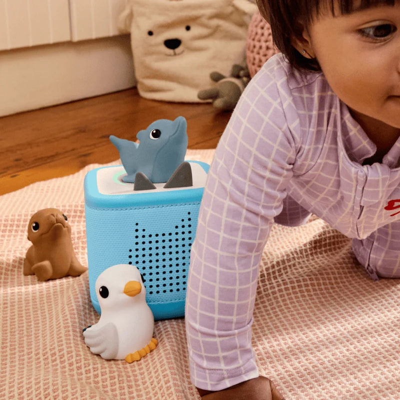 A baby crawling on a pink textured blanket next to a light blue toy basket filled with bath toys, including a blue dolphin, a white duck, and a brown seal in a home setting.