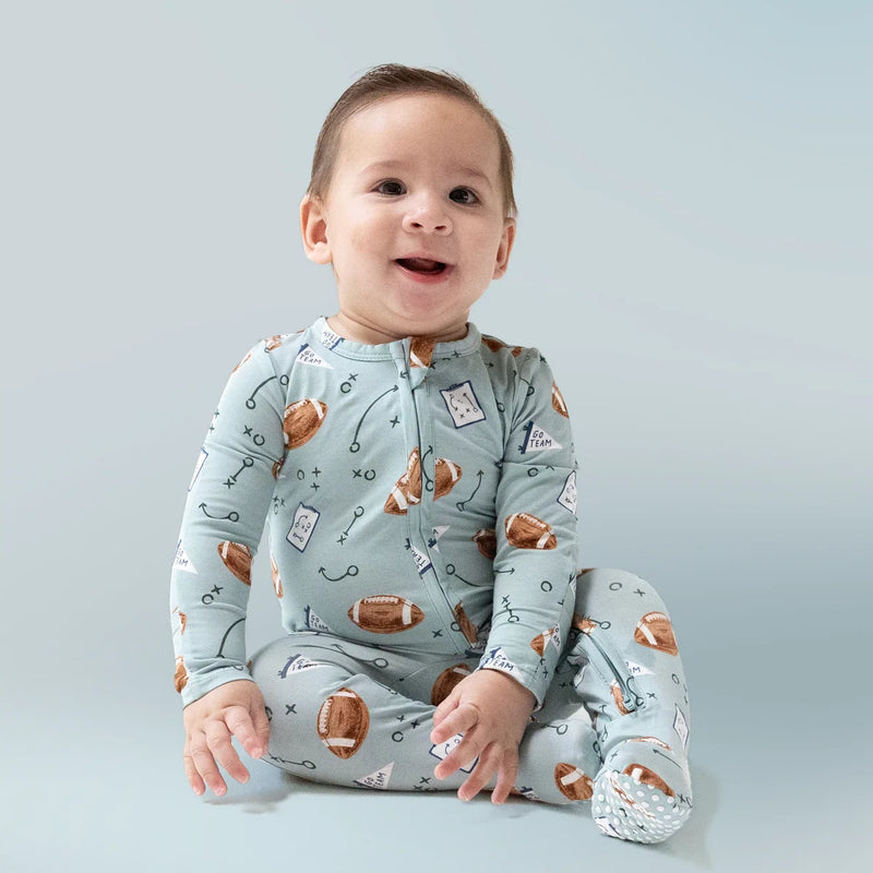 Smiling baby sitting upright wearing the same football-print romper against a soft blue backdrop.