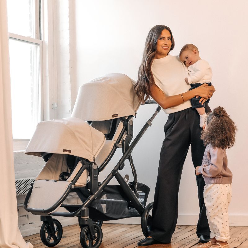 Woman holding a baby and standing next to a stroller in a well-lit room.