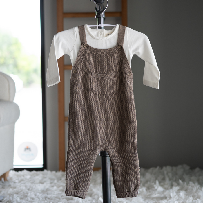 Brown knitted overalls on a hanger with a white shirt underneath, against a neutral background.