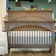 A wooden crib with an arched headboard and guard rails, shown in a furniture showroom with a white mattress inside. The crib has a weathered gray-brown wood finish.