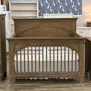 A rustic brown wooden crib with an arched headboard and guard rails, shown in a furniture showroom with a mattress inside.