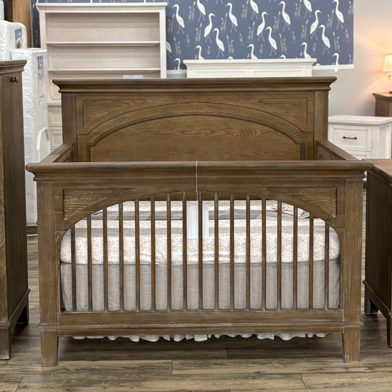 A rustic brown wooden crib with an arched headboard and guard rails, shown in a furniture showroom with a mattress inside.
