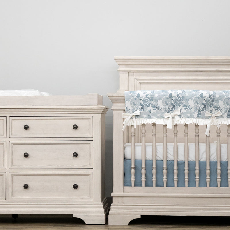 White nursery furniture set with crib and dresser against a light gray wall.